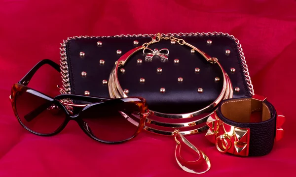Fashionable handbag and golden jewelry, glasses on red background.