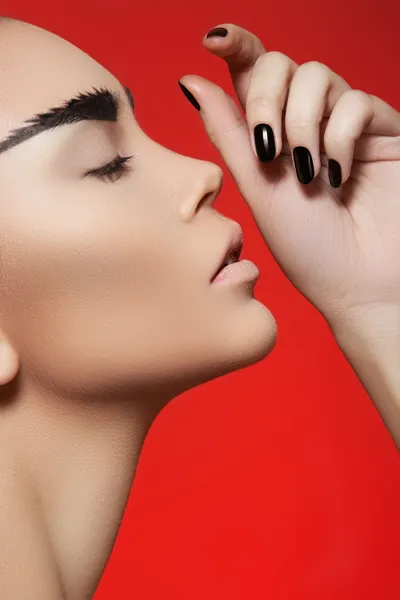 Fashion style, manicure, cosmetics and make-up. Profile portrait of beautiful woman with creative strong eyebrows makeup, clean skin, cheekbones and dark manicure on bright red background