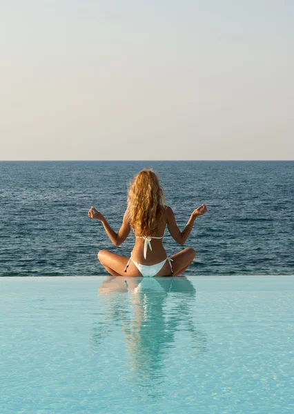 Rear view of woman in white bikini meditating on infinity pool