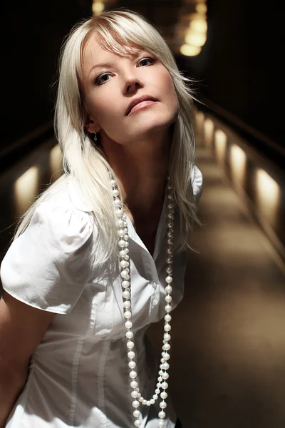 Young blonde model girl close up portrait in her early 30s or 20s looking straight in a white shirt with a white pearls with flare lights on the background in a tunnel
