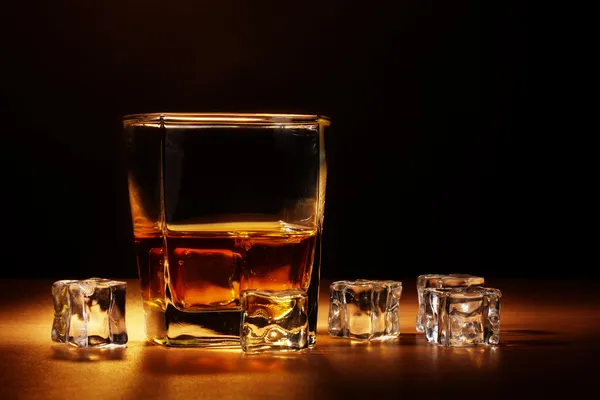 Glass of scotch whiskey and ice on wooden table on brown background