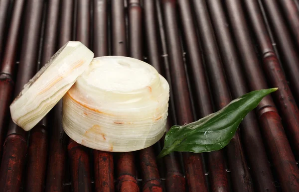 Opened jar of cream with fresh green leaf on bamboo mat with water droplets