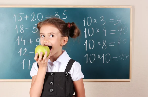 Beautiful little girl with apple standing near blackboard in classroom