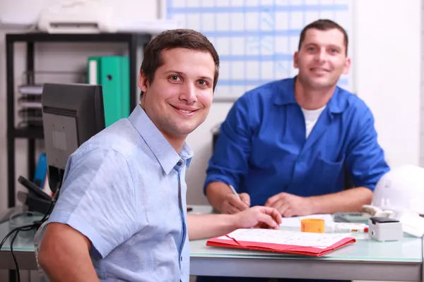 Two male colleagues sat in an office smiling and watching the camera
