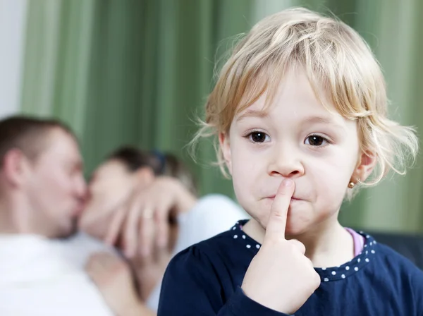 Blonde little girl making silence sign while mom and dad kissing in the background