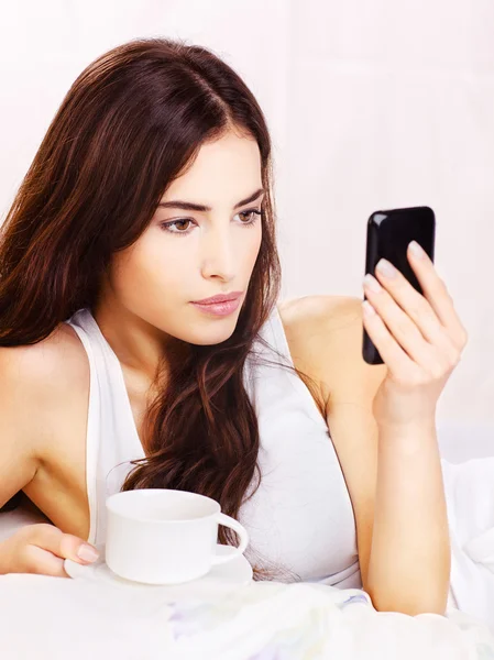 Woman making phone call and holding cup of coffee in bed