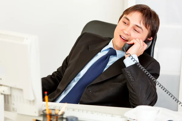 Pleased modern businessman sitting at office desk and talking on phone
