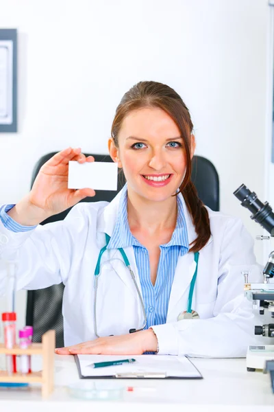 Smiling doctor woman sitting at office table and holding blank business car