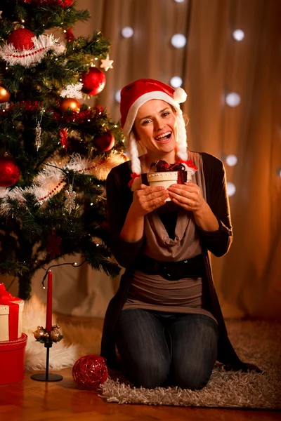 Happy young woman near Christmas tree hugging present box