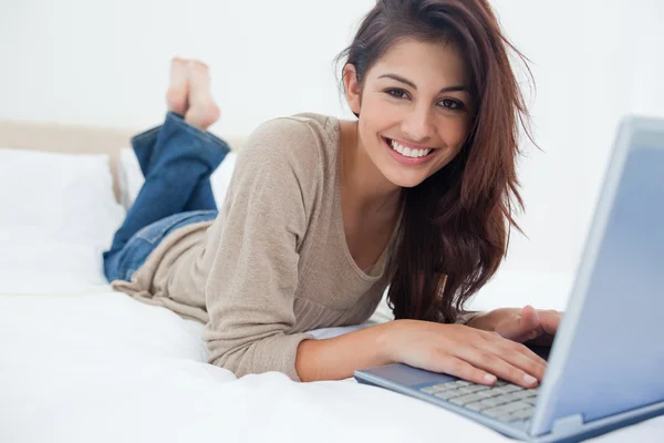 Woman smiling as she lies on the bed with her laptop in front of