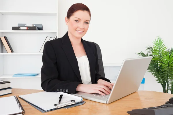 Young beautiful red-haired female in suit typing on her laptop