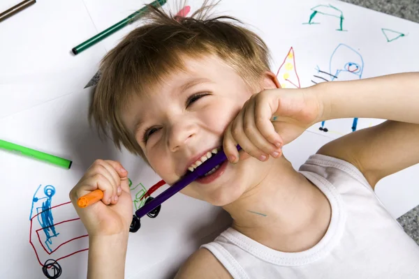 Laughing boy lying on his drawing with felt-tip pen in his teeth