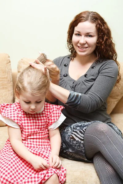 Мати і маленька донька сидять на дивані. Жінка розчісує волосся дочки — стокове фото