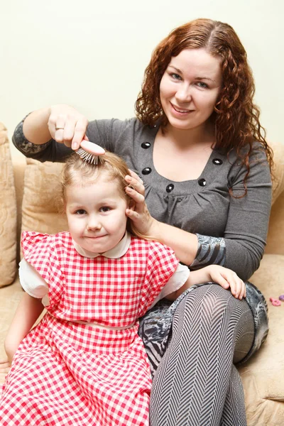 Mother and little daughter sitting on couch. Woman combing daughter hair — Stock Photo, Image