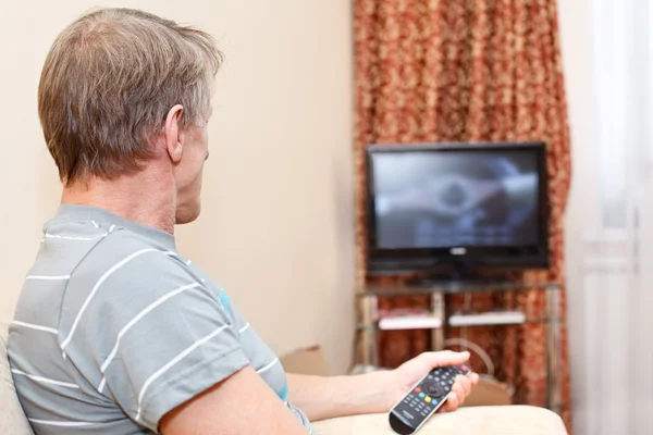 Senior man with remote control sitting on couch and looking at tv set — Stock Photo, Image
