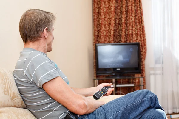 Senior man with remote control sitting on couch and looking at tv set — Stock Photo, Image