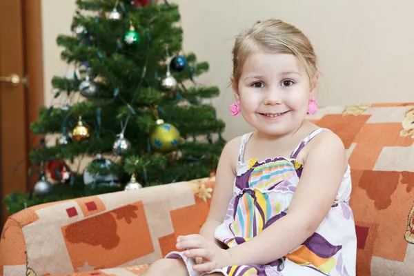 Pequena menina sorridente sentada no sofá atrás da árvore de Natal — Fotografia de Stock