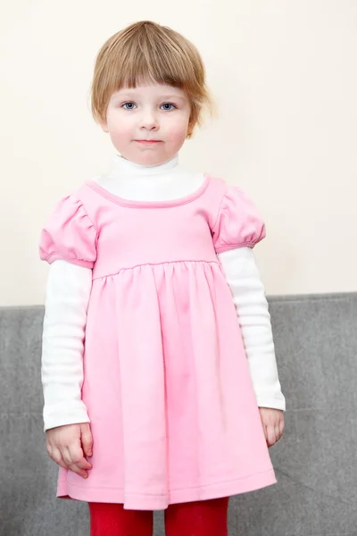 Portrét malé děvče v růžových šatech, stojící na gauči a hledá — Stock fotografie