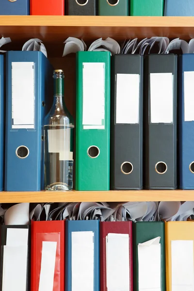 Glass bottle with alcohol hidden between file binders on shelves — Stock Photo, Image