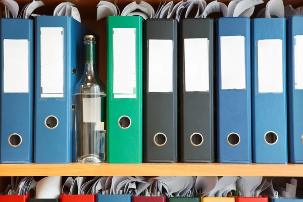 शेल्फ पर फ़ाइल बाइंडरों के बीच छिपे हुए शराब के साथ ग्लास बोतल — स्टॉक फ़ोटो, इमेज
