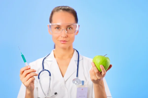 Young woman doctor injecting green apple with syringe on blue — Stock Photo, Image