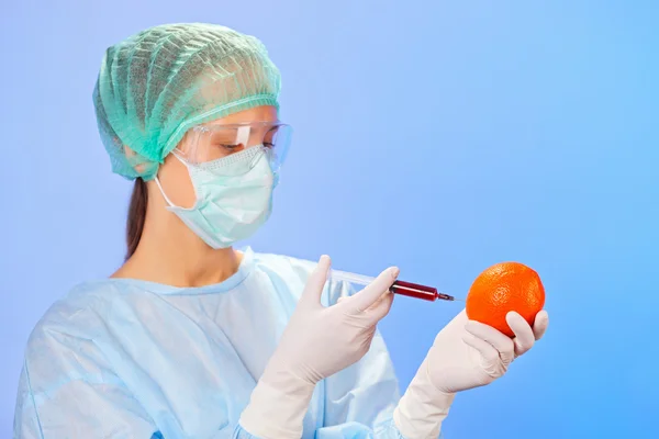 Young woman doctor injecting orange fruit with syringe on blue — Stock Photo, Image