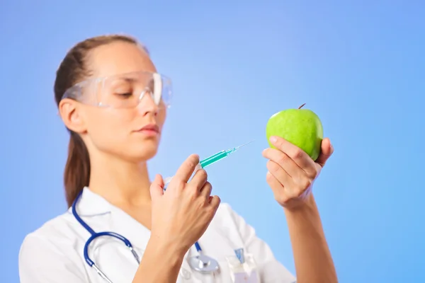 Young woman doctor injecting green apple with syringe on blue — Stock Photo, Image