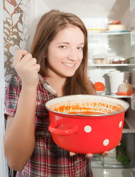 Девушка ест суп из кастрюли рядом с холодильником — стоковое фото