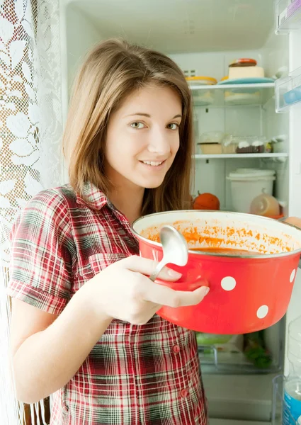 Dziewczynka jedzenie zupy z patelni w pobliżu lodówka — Zdjęcie stockowe