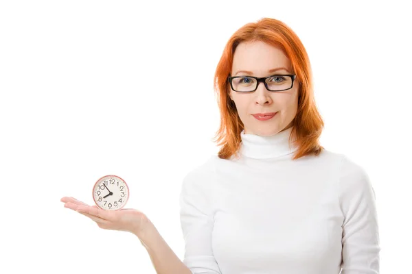 Una bella ragazza con i capelli rossi che indossa gli occhiali indica l'orologio su uno sfondo bianco . — Foto Stock