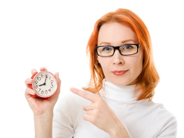Una bella ragazza con i capelli rossi con gli occhiali punta il dito contro l'orologio su uno sfondo bianco . — Foto Stock