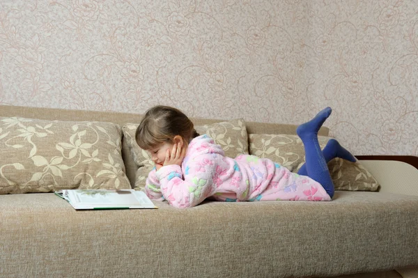 Маленька дівчинка читає книгу, що лежить на дивані — стокове фото