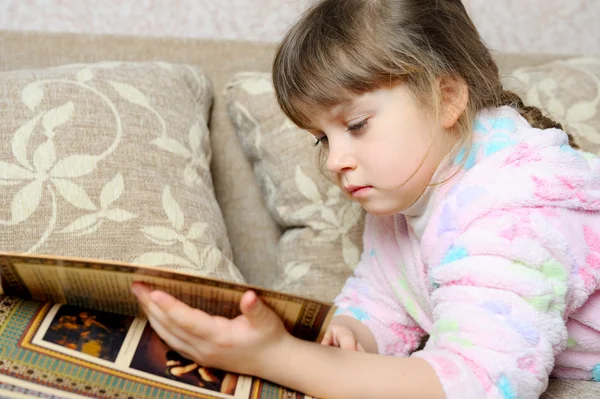 La bambina legge il libro sdraiata su un divano — Foto Stock