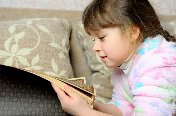 La bambina legge il libro sdraiata su un divano — Foto Stock