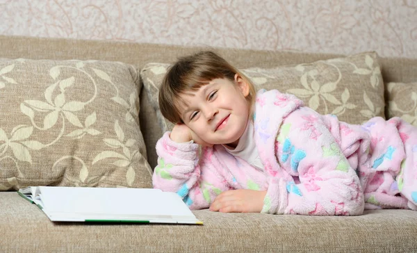 Das kleine Mädchen liest das Buch auf dem Sofa liegend — Stockfoto