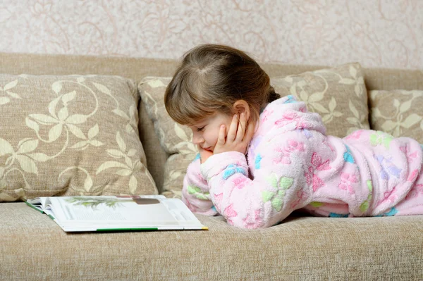 Das kleine Mädchen liest das Buch auf dem Sofa liegend — Stockfoto