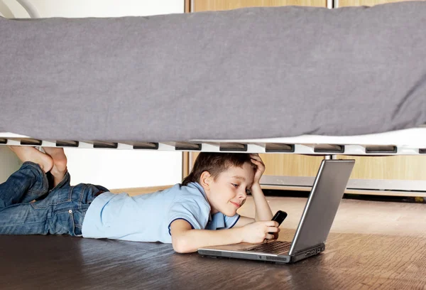 Dizüstü bilgisayar ve telefon yatağın altında çocuk — Stok fotoğraf