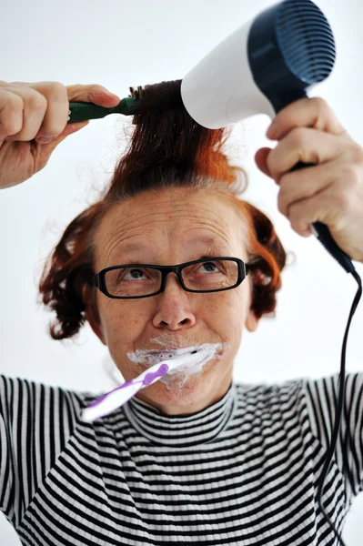 Senior woman brushing teeth and drying hair — Stock Photo, Image