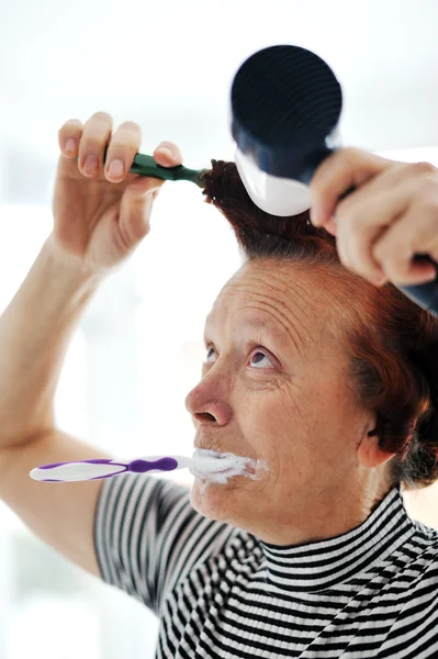 Femme âgée séchant les cheveux avec ventilateur et brossant les dents — Photo