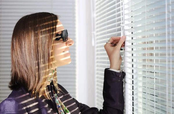 Jonge brunette vrouw in het zonlicht op zoek via jalousie op venster — Stockfoto