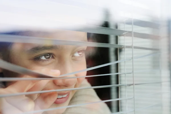 Porträt eines kleinen Mädchens, das mit Jalousie durch Fenster guckt — Stockfoto