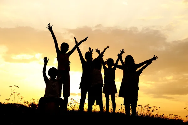 Silueta, skupina šťastných dětí hraje na louce, západ slunce, léto — Stock fotografie