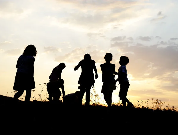 Silhouette, grupp av glada barn spelar på ängen, solnedgång, sommartid Royaltyfria Stockfoton