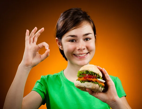 लड़की बड़ा हैम्बर्गर खा रही है जो ठीक संकेत दिखाती है — स्टॉक फ़ोटो, इमेज