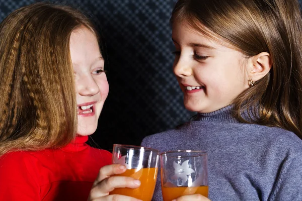 Two little girls holding glasses and drinking orange fruit — Stock Photo, Image