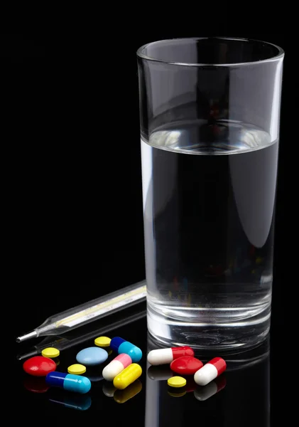 Pastillas, termómetro y vaso de agua sobre fondo negro — Foto de Stock