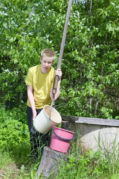 Der Junge, der Teenager, gießt Wasser in einen Eimer aus einem Brunnen — Stockfoto