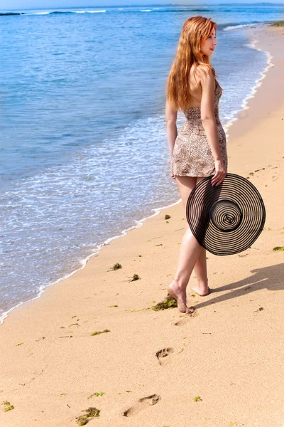 Young graceful woman goes on coast of ocean with straw hat in hands — Stock Photo, Image