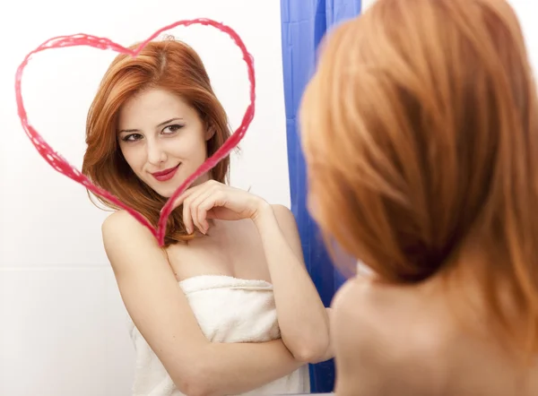 Rödhårig flicka nära spegel med hjärtat på det i badrum. — Stockfoto