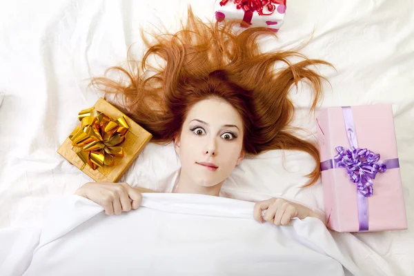 Έκπληκτος κοκκινομάλλα κοπέλα στο κρεβάτι με δώρα. — Φωτογραφία Αρχείου
